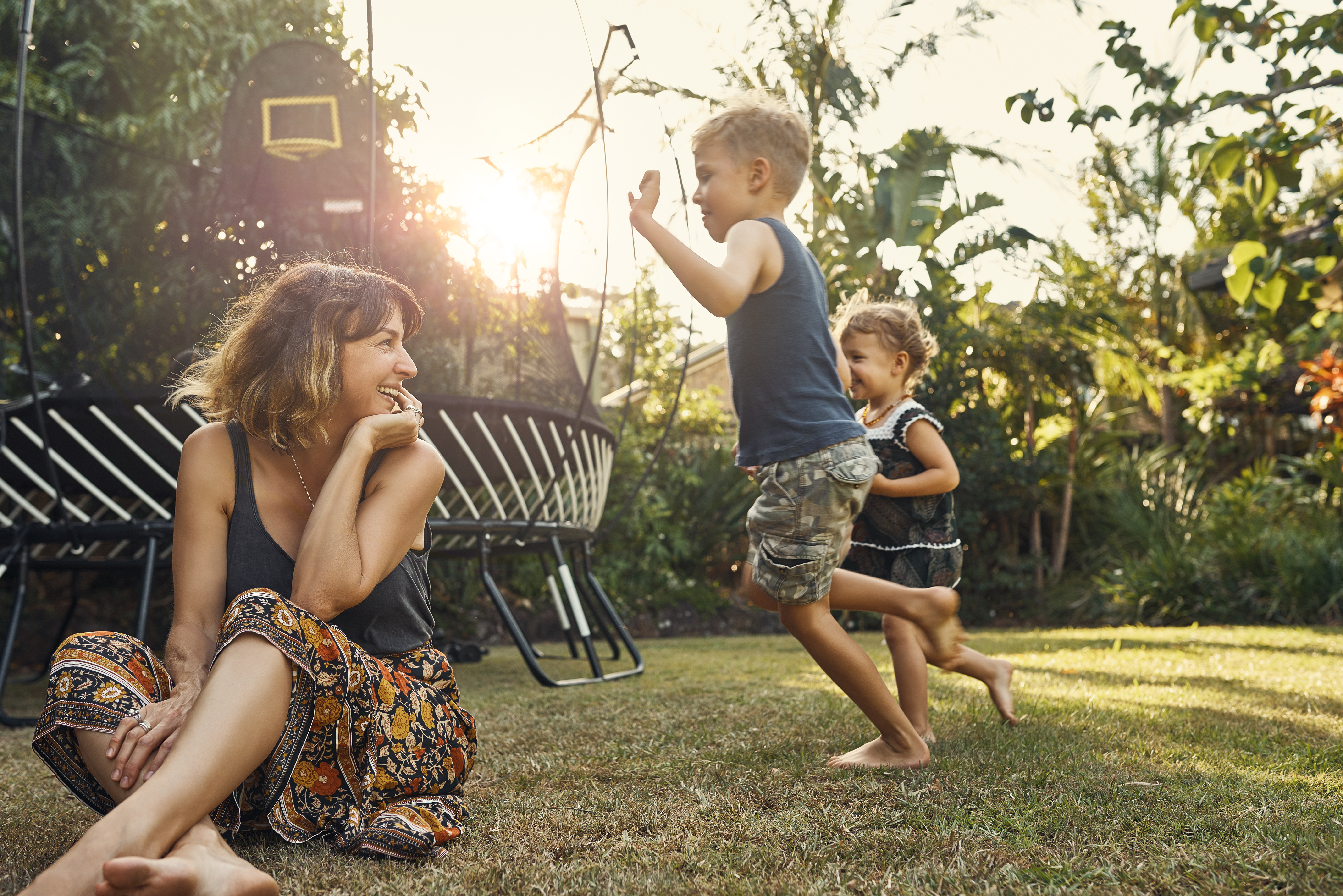 Kids playing in the backyard with mum sitting in the front, smiling